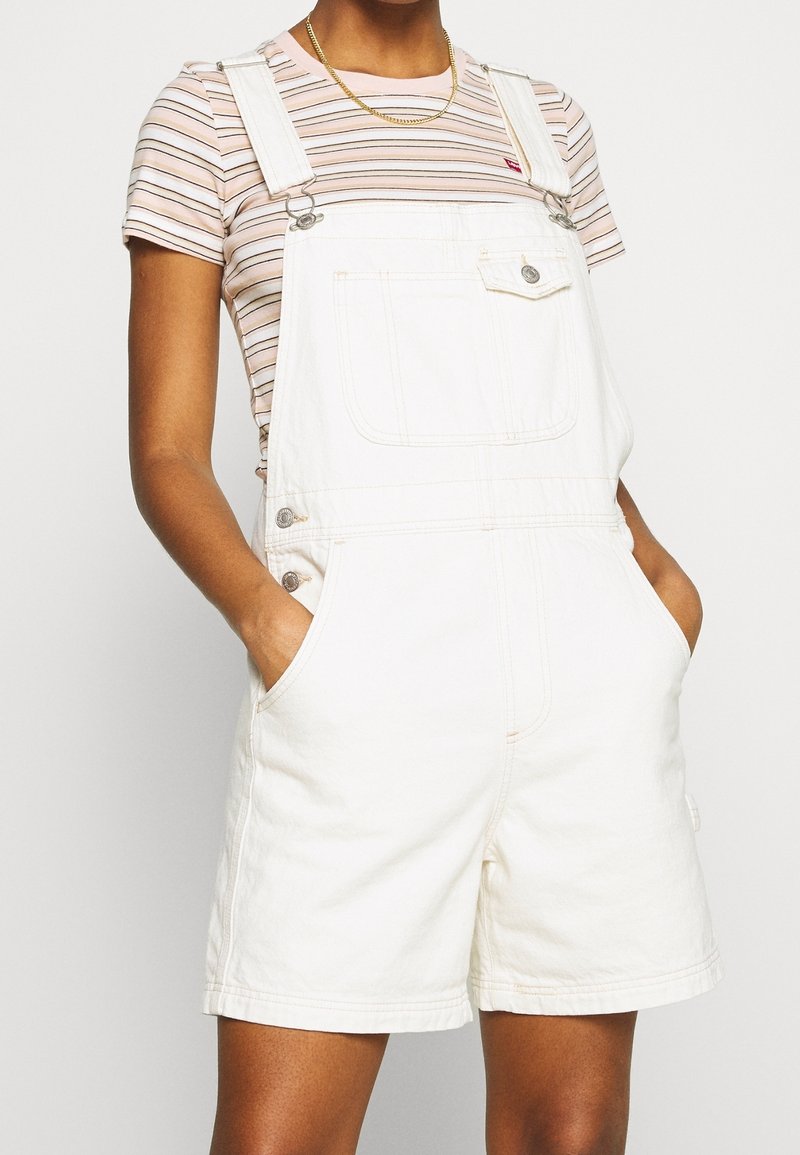 Personne portant une salopette courte en denim blanc avec une poche avant sur un t-shirt à manches courtes rose et blanc à rayures, mains dans les poches.