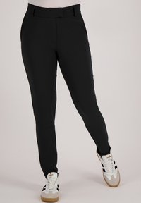 Zwarte slim-fit broek met een gladde textuur, voorzien van een middelhoge tailleband en subtiele plooien aan de voorkant, gecombineerd met witte sneakers.