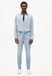 Chemise à rayures bleu clair, t-shirt noir en dessous, jeans clair, et baskets blanches. Le mannequin se tient contre un fond blanc uni.