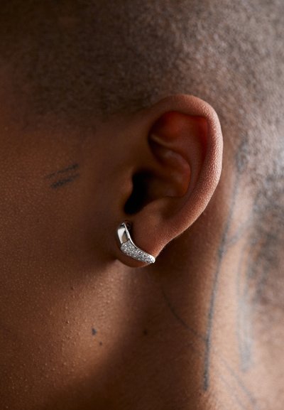 Boucle d'oreille en argent au design courbé, ornée d'une section de pierres brillantes, attachée à l'oreille d'une personne aux cheveux courts.