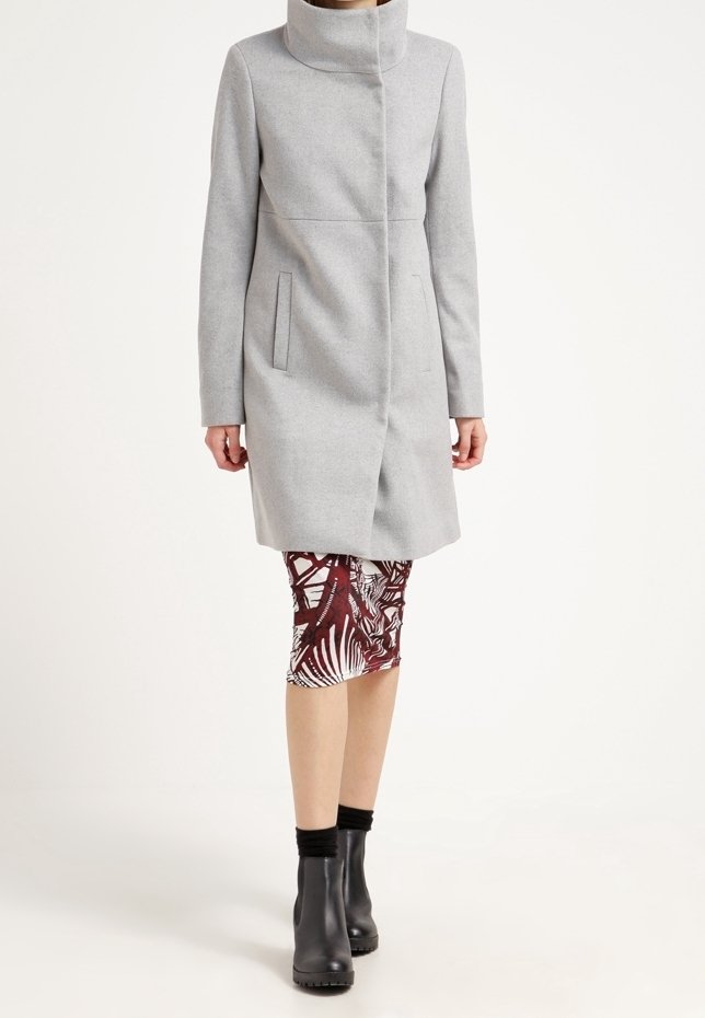 Femme portant un manteau gris clair à col montant, une jupe à motifs longueur genoux, des bottines noires et des chaussettes noires.