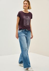Lilla bomulds t-shirt med et grafisk design i midten, parret med lyseblå brede jeans og hvide sneakers. Glat tekstur.