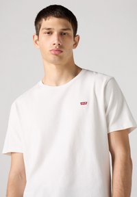 Witte katoenen T-shirt met ronde hals, korte mouwen en een klein rood Levi's-logo op de linkerborst. Gladde textuur, standaard pasvorm.