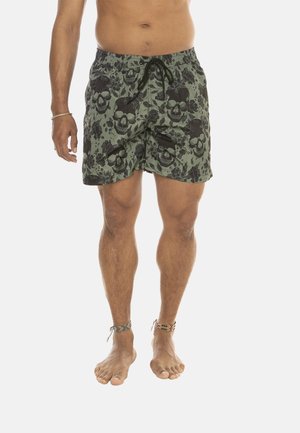 Homme portant un short de bain vert avec des motifs noirs de crânes et floraux, pieds nus avec des bracelets de cheville tissés, debout devant un fond blanc.