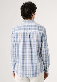 Muž s tmavými kadeřavými vlasy, oblečený v dlouhém rukávu s modro-bílo-zeleným kostkovaným vzorem, stojí odvrácen od jasného pozadí.