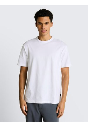 Jonge man met krullend haar, gekleed in een effen wit T-shirt met korte mouwen en een grijze broek, staand tegen een lichtgrijze achtergrond.