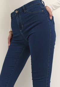 Tamno plavi skinny traperice s visokim strukom, kontrastnim šavovima i pet džepova, s metalnim dugmetom za zakopčavanje.
