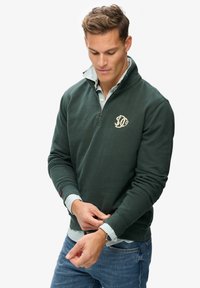Homme ajustant le poignet d'un sweat-shirt vert forêt à fermeture éclair quart, porté sur une chemise rayée à col, portant un jean bleu et une montre-bracelet, sur fond blanc.