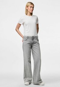 Sive široke traperice s klasičnim pet džepova, uparene s običnom bijelom pamučnom majicom. Model stoji s rukama na bokovima.