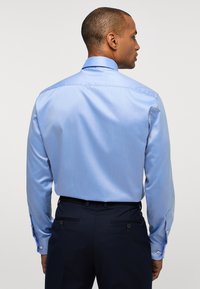 Camicia a maniche lunghe azzurro chiaro con una texture liscia e colletto classico; abbinata a pantaloni scuri con due tasche posteriori e bottoni.