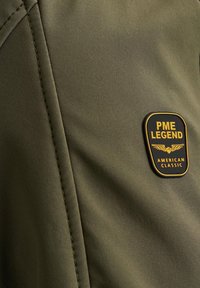 Tkanina maslinaste boje s glatkom teksturom, koja uključuje crnu i zlatnu oznaku PME Legend s natpisom "AMERICAN CLASSIC." Vidljivi detalj šava.