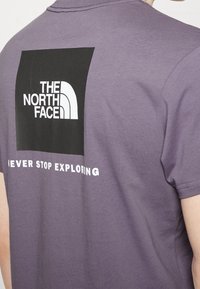 Fialové bavlněné tričko s černým čtvercovým potiskem na zadní straně, na kterém je bíle napsáno "THE NORTH FACE" a "NEVER STOP EXPLORING".