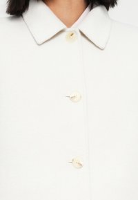 Λευκό μάλλινο παλτό με κλασικό γιακά, τέσσερα μεγάλα μπεζ κουμπιά και απαλή υφή. Καθαρές γραμμές χωρίς ορατά μοτίβα ή διακοσμητικά στοιχεία.