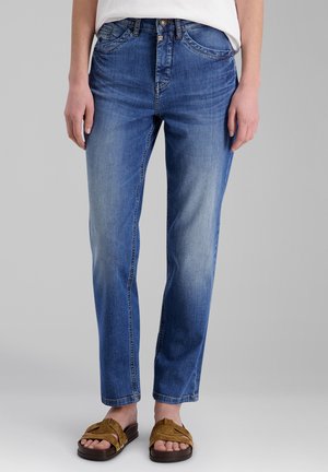 Person trägt blaue, verwaschene, gerade geschnittene Jeans mit weißem Oberteil und braunen offenen Sandalen vor einem schlichten grauen Hintergrund.