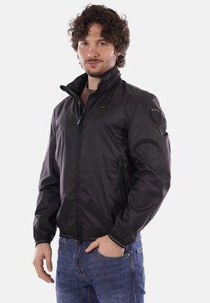 Uomo con capelli ricci che indossa una giacca nera con cerniera e collo alto e jeans blu, in piedi con una mano in tasca.