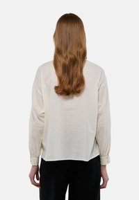 Blouse couleur crème en tissu léger, avec des manches longues, des poignets froncés et une garniture décorative le long des manches. Texture lisse.