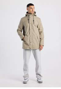 Beige waterproof jacket with a hood, front buttons, and drawstrings. Paired with light gray sweatpants and athletic shoes.