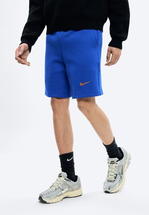 Muž oblečený v modrých šortkách Nike, čiernych ponožkách Nike a strieborných teniskách Nike, stojaci so skríženými nohami na bielom pozadí.