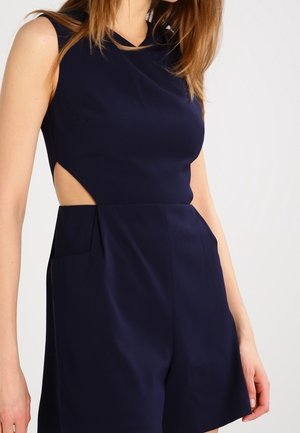 Laivastonsininen, hihaton haalari, jossa on korkea pääntie, sivuleikkaukset ja laskosyksityiskohdat shortseissa lisätyn tekstuurin ja rakenteen aikaansaamiseksi.