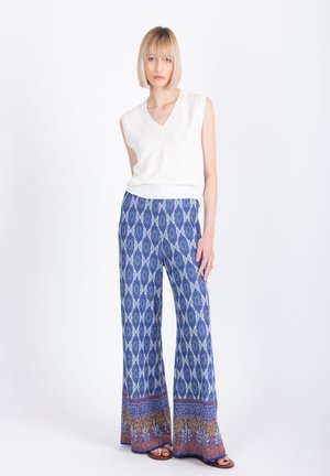 Donna in piedi che indossa un top bianco senza maniche con scollo a V e pantaloni ampi a fantasia blu con sandali, su sfondo bianco.