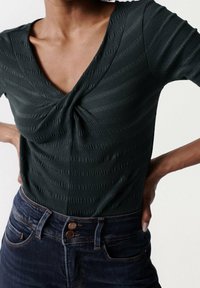 Dunkelgrüner gerippter Strickbody mit tiefem V-Ausschnitt und verdrehtem Frontdetail, kombiniert mit hoch geschnittenen dunklen Jeans.