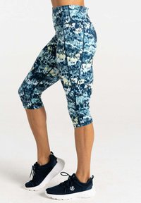 Hochgeschnittene Capri-Leggings in einem blauen, grünen und gelben abstrakten Muster mit glatter Textur; getragen mit schwarzen Sportschuhen.