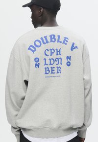 Hanorac gri cu text grafic albastru pe spate, având inscripția "DOUBLE A CPH LDN BER 2020" în diferite stiluri și dimensiuni de font.