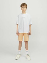 Baltas oversize T-shirt su juodu logotipu, derinamas su šviesiai oranžiniais šortais, kuriuose yra tekstas. Balti sportiniai bateliai su žaliais akcentais ir balti keturkampiai kojinės.
