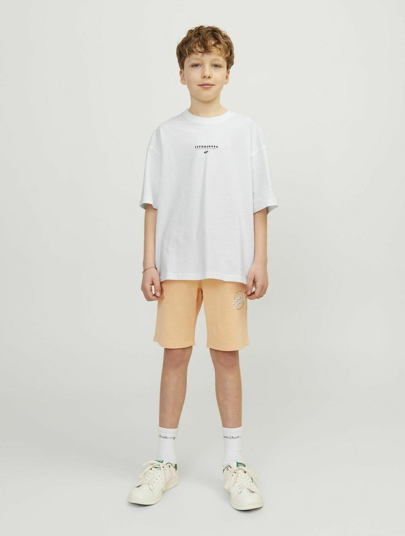 Baltas oversize T-shirt su juodu logotipu, derinamas su šviesiai oranžiniais šortais, kuriuose yra tekstas. Balti sportiniai bateliai su žaliais akcentais ir balti keturkampiai kojinės.