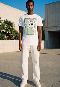 Fiatal férfi fehér Bugs Bunny mintás pólóban, fehér nadrágban és fehér sportcipőben, betonépületek és pálmák közelében, szabadban áll.