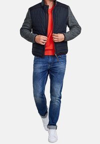 Muž v černé prošívané vestě přes šedé dlouhé rukávy a červenou košili, v modrých vyhrnutých džínách a bílých teniskách, zipuje vestu.