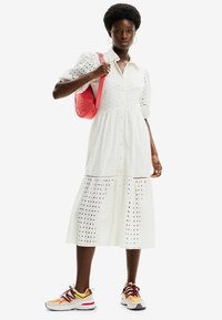 Abito midi bianco in pizzo con maniche a palloncino corte, chiusura con bottoni e design a strati. Abbinato a sneakers sportive multicolori e a una borsa rosa.