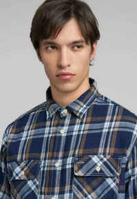 Camisa de franela con un patrón de cuadros en azul, marrón y blanco, cuello abotonado y dos bolsillos en el pecho con botones. Textura de tela suave.