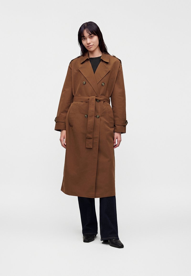 Femme debout portant un trench-coat marron à ceinture sur un pantalon noir et des chaussures noires sur un fond clair uni.
