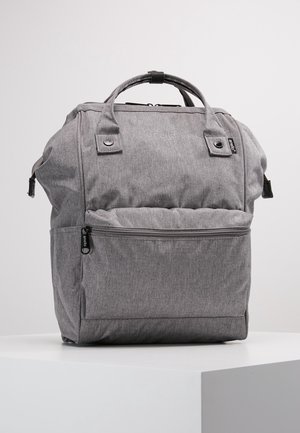 Sac à dos en tissu gris avec poignées supérieures, poche avant zippée et tirettes de fermeture éclair noires, posé sur un socle blanc sur fond uni.