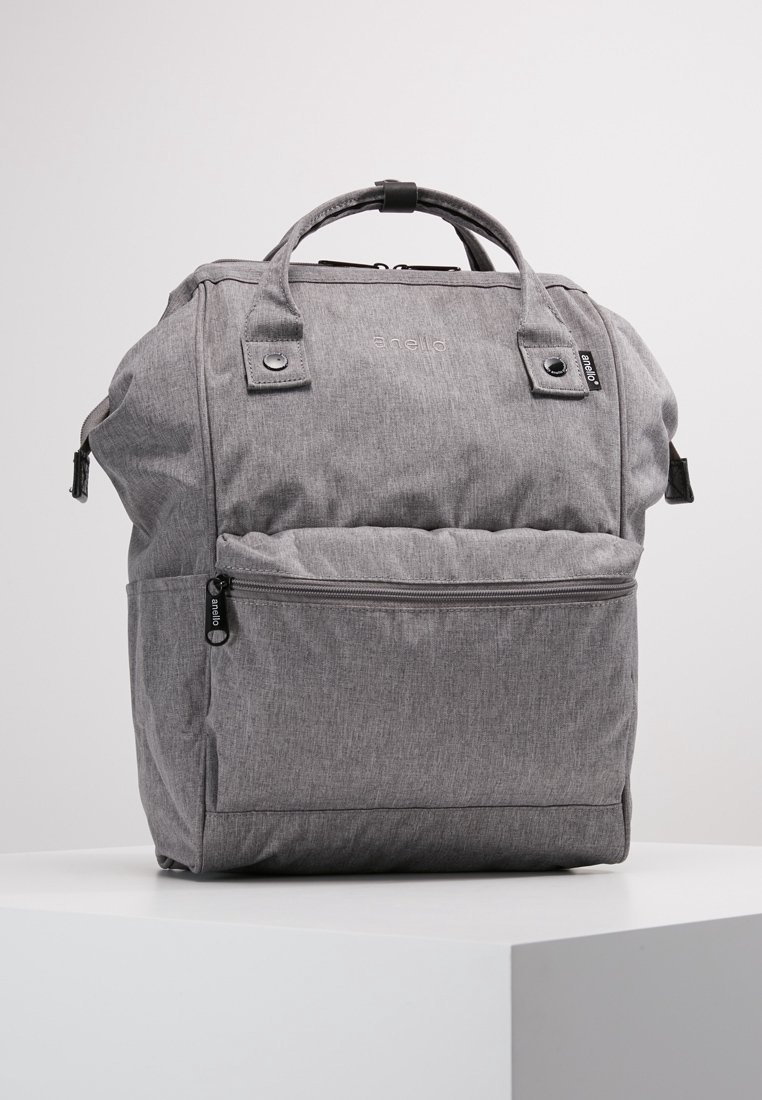 Sac à dos en tissu gris avec poignées supérieures, poche avant zippée et tirettes de fermeture éclair noires, posé sur un socle blanc sur fond uni.