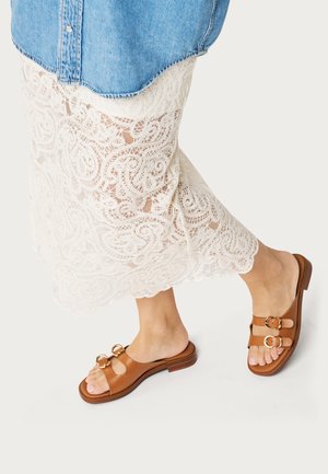 Pies con sandalias marrones de doble tira con hebillas doradas, vistiendo una falda blanca de encaje y una camisa azul de mezclilla, pisando sobre un fondo claro.