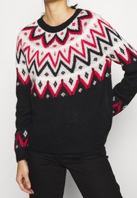 Strickpullover mit rundem Ausschnitt, der ein geometrisches Muster in Rot, Schwarz und Weiß aufweist. Lange Ärmel mit gemusterten Bündchen.