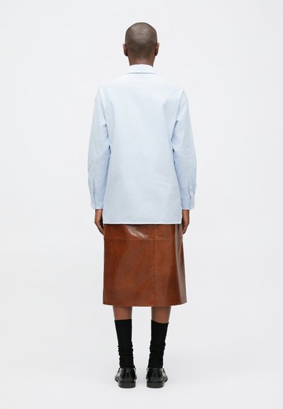 Asmuo stovi atsukęs nugarą, vilkintis šviesiai mėlyną ilgomis rankovėmis marškinį, rudą odinę sijoną, juodas kojines ir juodus batus ant balto fono.