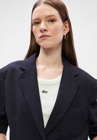 Blazer bleu marine au design structuré et à une seule poche, porté sur un haut de couleur claire avec un petit logo vert Lacoste.