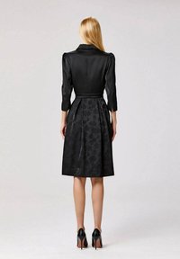 Femme aux longs cheveux blonds portant une robe noire mi-longue avec une jupe à motif floral et des manches trois-quarts, associée à des talons hauts noirs, de dos.