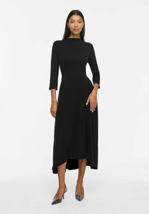 Le mannequin se tient devant un fond blanc, portant une robe midi noire à manches trois-quarts et des escarpins noirs à bout pointu.