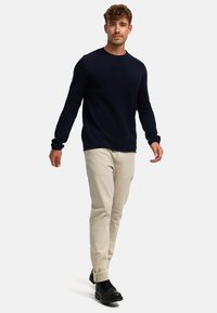 Marineblauer Pullover aus strukturiertem Strickstoff, kombiniert mit hellbeigen Chinos und schwarzen Schnürstiefeln. Das Model läuft vor einem schlichten weißen Hintergrund.