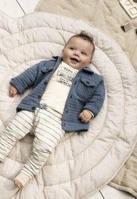 Blau gestepptes Jacket mit Taschen, cremefarbenes Hemd mit Kameragrafik und cremefarbene gestreifte Hose. Liegt auf einem runden gepolsterten Matratze.