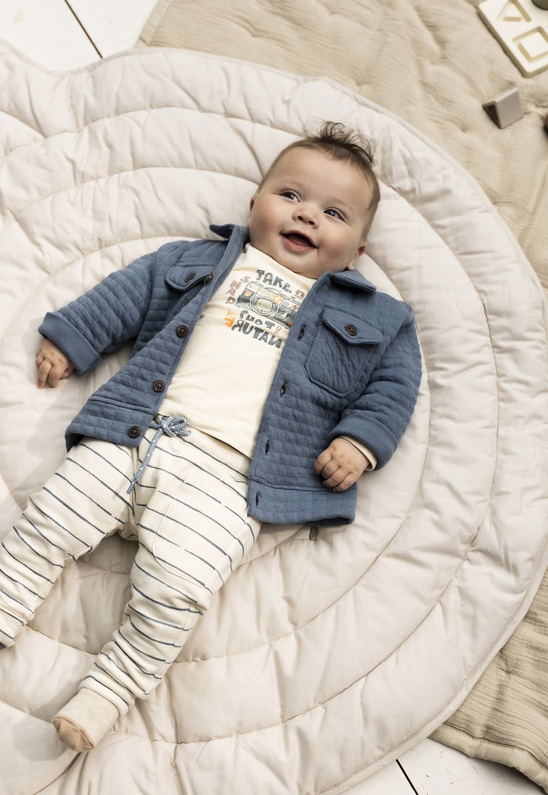 Blau gestepptes Jacket mit Taschen, cremefarbenes Hemd mit Kameragrafik und cremefarbene gestreifte Hose. Liegt auf einem runden gepolsterten Matratze.