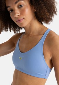 Sujetador deportivo azul claro hecho de tela elástica con escote redondo, tirantes anchos y un pequeño logo de corazón verde en el frente.