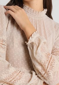 Helle beige Bluse aus strukturiertem Stoff mit zarten Punkten, hohem Rüschenausschnitt und Spitzenakzenten an den Ärmeln.