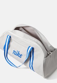 Saco de duffel cinza com pegas azul e brancas listradas, logótipo "nike" bordado em azul, apresentando uma abertura com fecho e textura suave.
