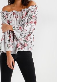 Bílá off-the-shoulder blůza s květinovým vzorem v růžové a zelené, s volnými rukávy a volánkovým výstřihem. V kombinaci s černými džínami.