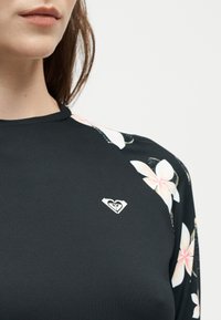 Gros plan d'une personne portant un haut noir avec des manches à motif floral et un petit logo blanc en forme de cœur sur la poitrine.